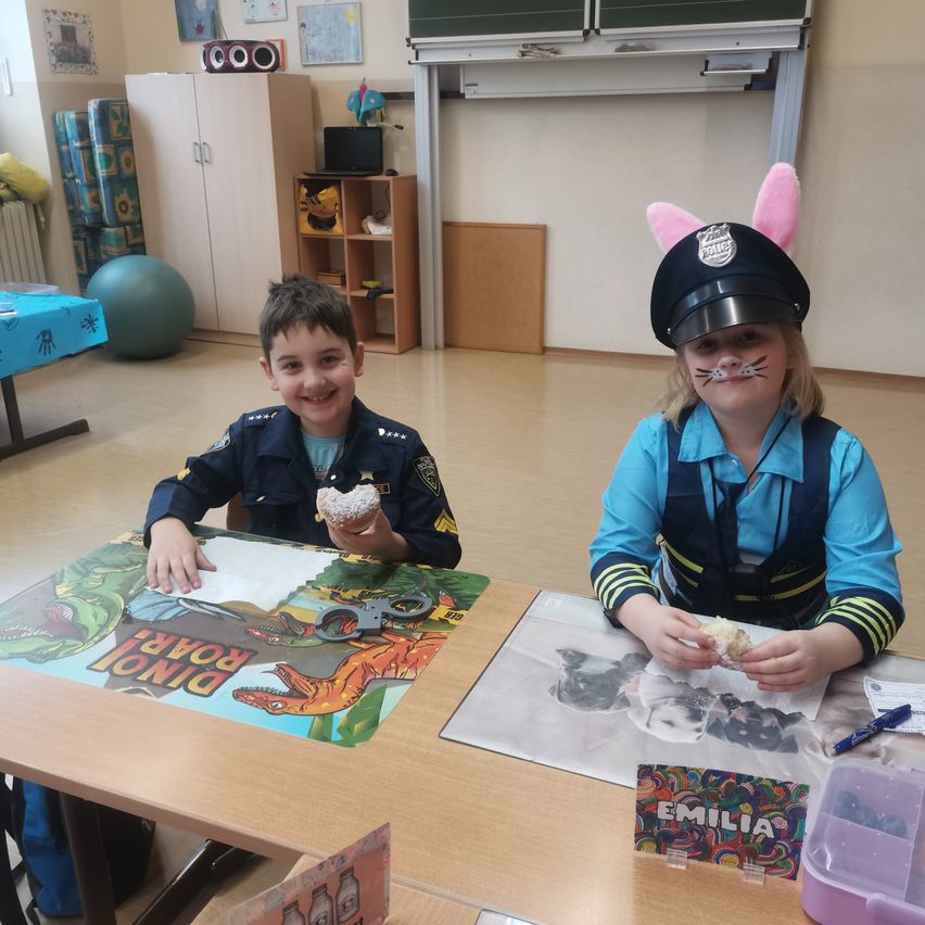 Zwei Kinder, die als Polizisten verkleidet sind, sitzen an einem Tisch, lächeln und essen Donuts. Der Tisch ist mit einem Dinosaurier-Thema-Matte bedeckt. Hinter ihnen ist ein Klassenzimmer mit verschiedenen Gegenständen an den Wänden und dem Boden.
