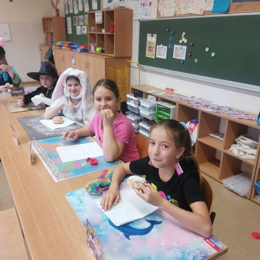 Eine Gruppe von Kindern sitzt in einem Klassenzimmer an einem Tisch und isst Donuts. Einige tragen Kostüme. Im Hintergrund befinden sich Papiere, Aufbewahrungsboxen und eine grüne Tafel mit Pinseln.