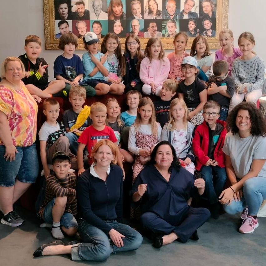 Eine Gruppe von Kindern und Erwachsenen posiert für ein Foto vor einer Collage von Porträts an der Wand.