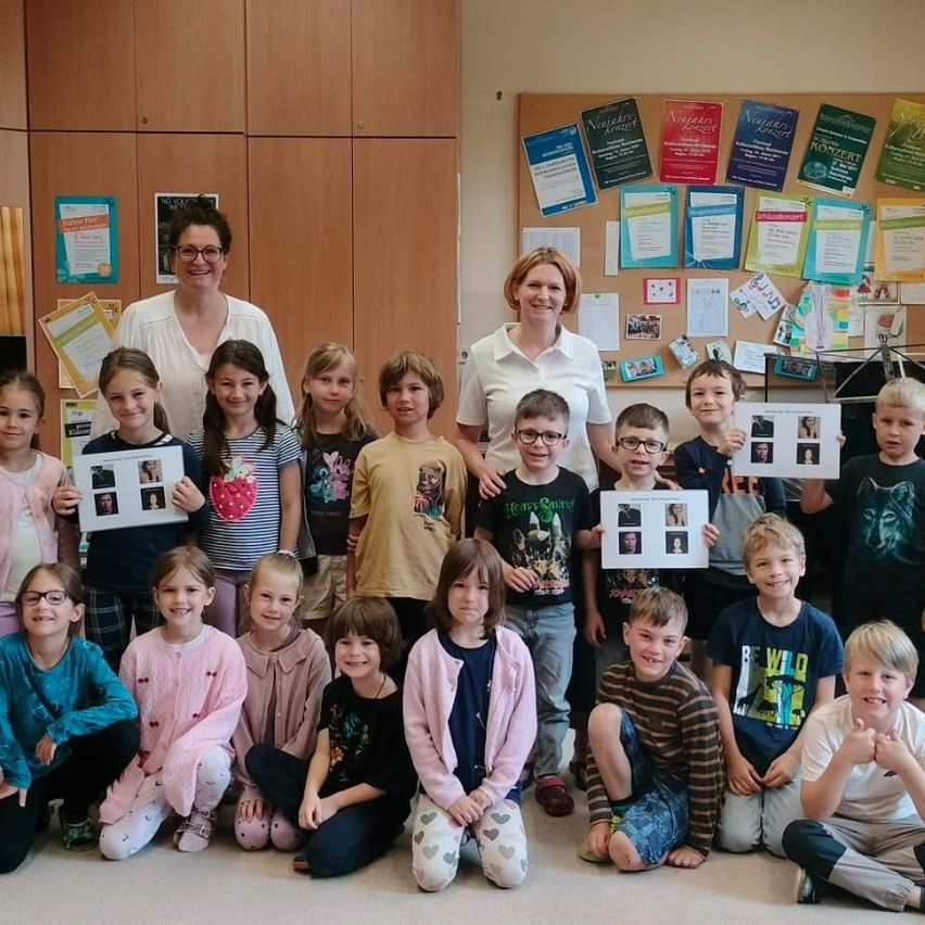 Klassenfoto mit Kindern, die Bilder hochhalten. Zwei lächelnde Frauen vorne. An der Wand ein Pinnwand mit Aushängen und Postern.