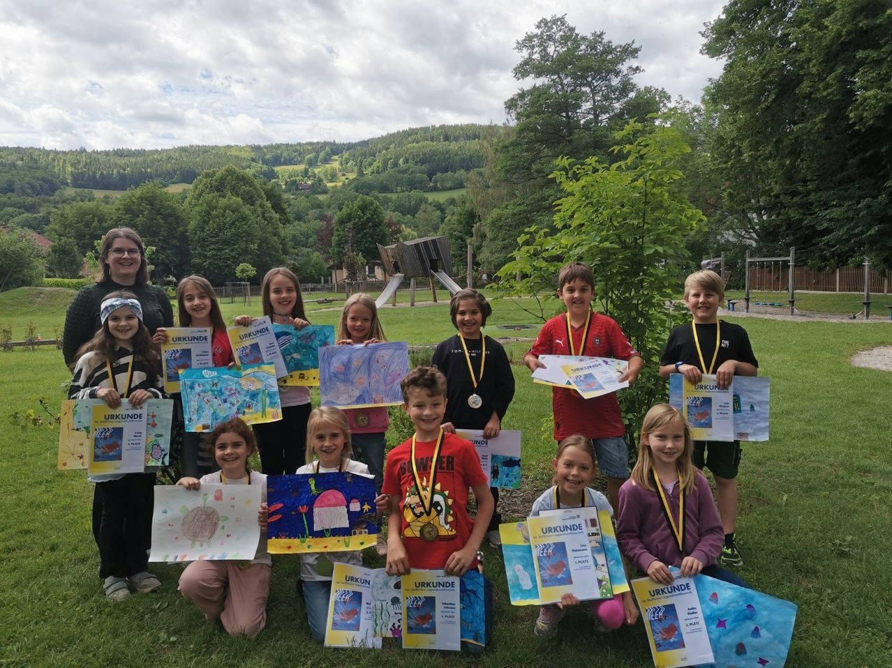 Eine Gruppe von Kindern mit Urkunden und Zeichnungen posiert für ein Foto im Park, mit einer Frau im Hintergrund und Bergen in der Ferne.