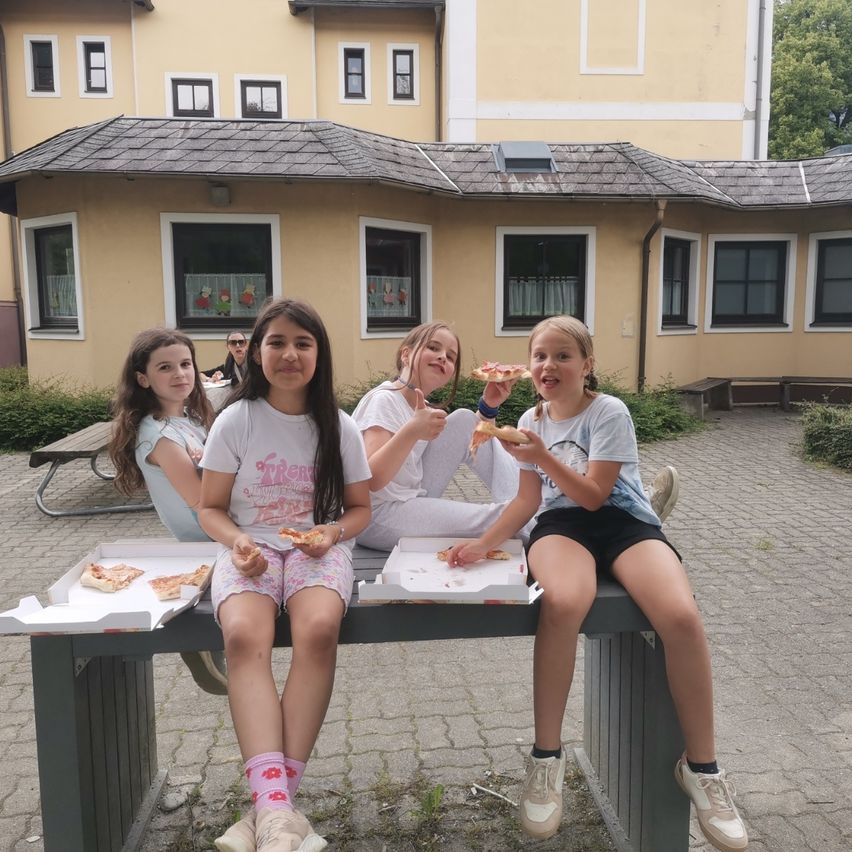 Fünf junge Mädchen sitzen auf einer Bank vor einem Gebäude und essen Pizza. Sie haben Pizzakartons vor sich. Das Gebäude hat mehrere Fenster und ein Dach. Eine Person sitzt an einem Picknicktisch.