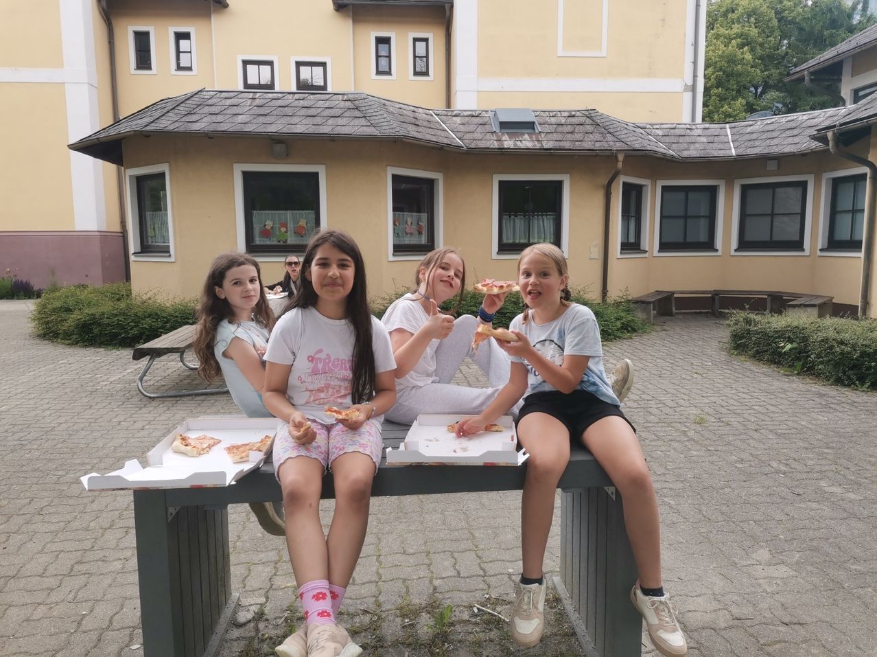 Fünf junge Mädchen sitzen auf einer Bank vor einem Gebäude und essen Pizza. Sie haben Pizzakartons vor sich. Das Gebäude hat mehrere Fenster und ein Dach. Eine Person sitzt an einem Picknicktisch.