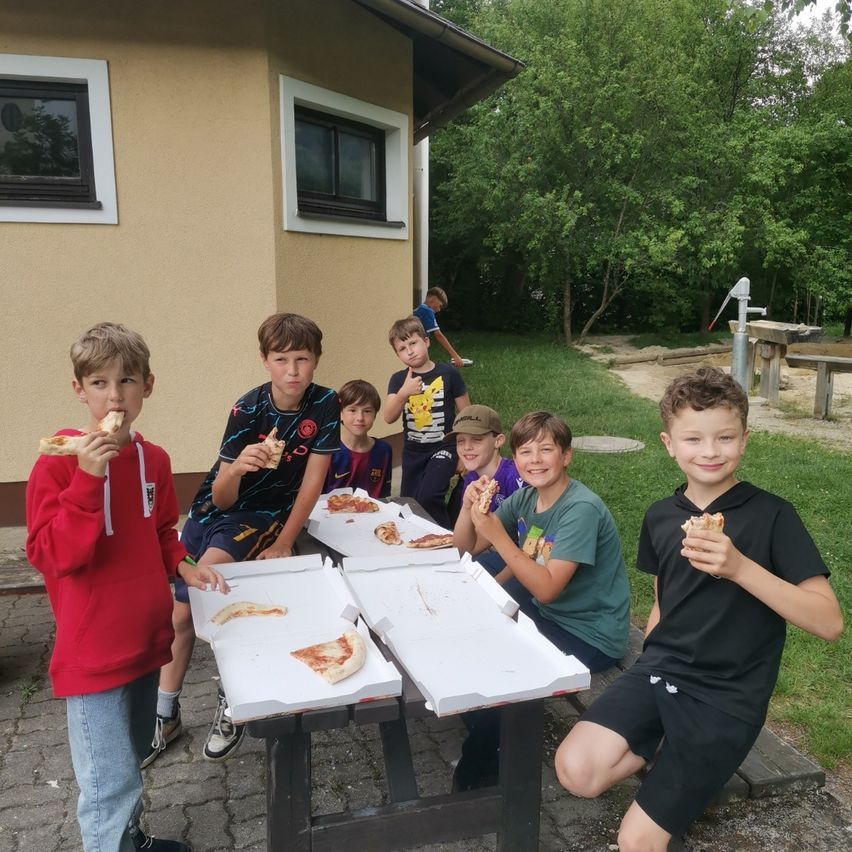 Eine Gruppe von Jungen sitzt an einem Picknicktisch und isst Pizza. Hinter ihnen steht ein Gebäude mit zwei Fenstern.