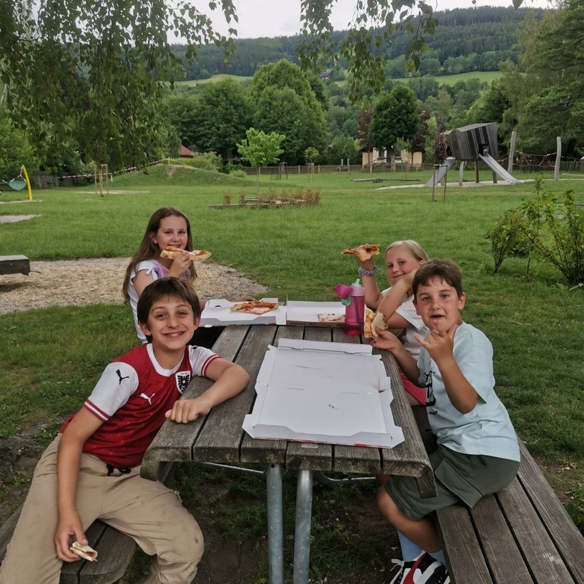 Fünf Kinder sitzen um einen Picknicktisch in einem Park und essen Pizza. Hinter ihnen befindet sich ein Rasenfeld mit Bäumen und einem Spielplatz.
