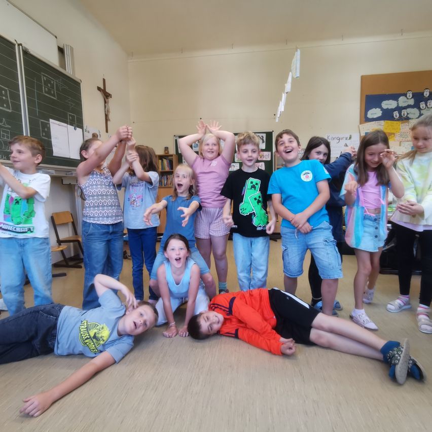 Eine Gruppe von Kindern in einem Klassenzimmer mit zwei Schülern auf dem Boden. Einige heben die Hände, während andere in die Kamera schauen. Der Raum hat eine weiße Wand und einen Holzboden.