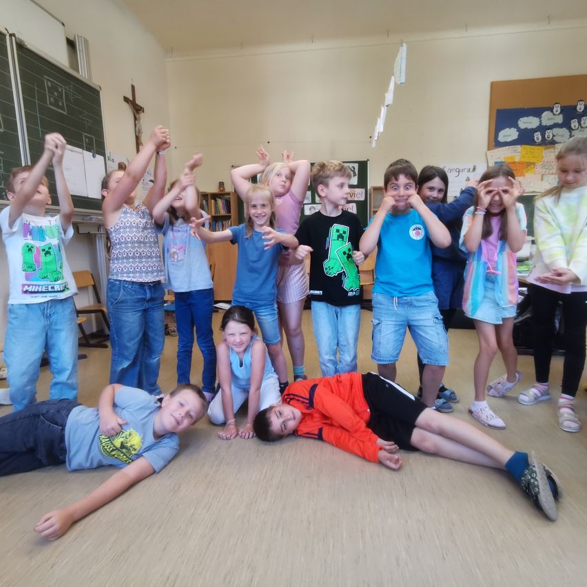 In einem Klassenzimmer posiert eine Gruppe von Kindern für ein Foto. Einige stehen mit erhobenen Händen, andere liegen auf dem Boden. Der Raum ist mit einer Tafel, einem Kreuz und verschiedenen Postern dekoriert.