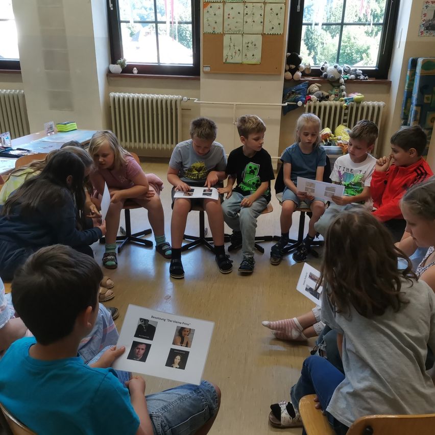 Ein Klassenzimmer gefüllt mit Kindern, die im Kreis sitzen. Sie halten Bücher und sehen sich Bilder von Menschen an. Einige tragen Sandalen, andere Turnschuhe. Ein Heizkörper und Spielzeug sind im Hintergrund.