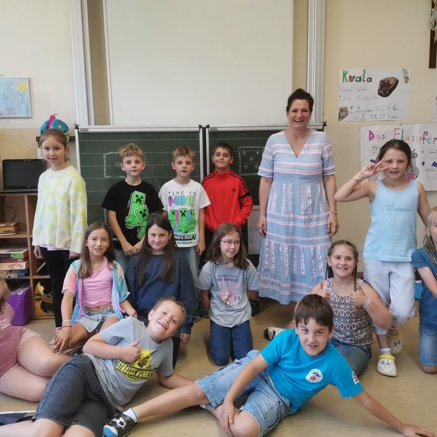 Eine Gruppe von Kindern und eine Lehrerin posieren für ein Foto im Klassenzimmer. Die Lehrerin lächelt und einige Kinder winken.