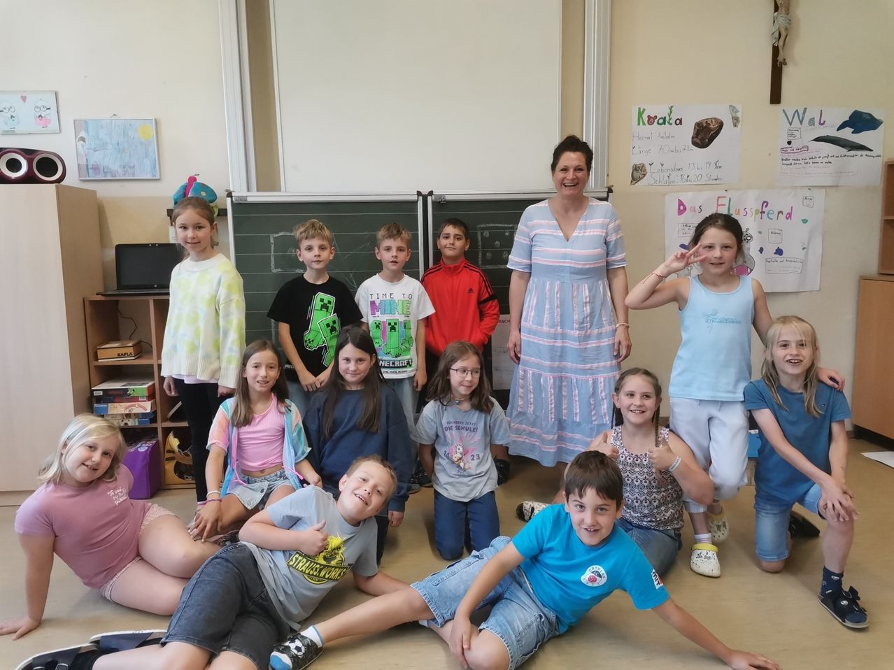 Eine Gruppe von Kindern und eine Lehrerin posieren für ein Foto im Klassenzimmer. Die Lehrerin lächelt und einige Kinder winken.