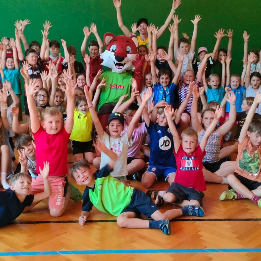 Gruppe von Kindern in einer Turnhalle mit einem Maskottchen, alle mit erhobenen Händen, einige sitzend und andere stehend, mit einer grünen Wand im Hintergrund.