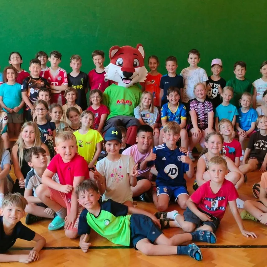 Eine Gruppe von Kindern und ein Maskottchen posieren für ein Foto vor einer grünen Wand. Das Maskottchen trägt ein rotes Fuchskostüm. Die Kinder tragen verschiedene Team-Trikots.