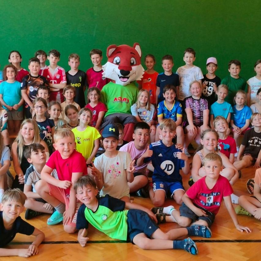 Eine Gruppe von Kindern und ein Maskottchen posieren für ein Foto vor einer grünen Wand. Das Maskottchen trägt ein rotes Fuchskostüm. Die Kinder tragen verschiedene Team-Trikots.