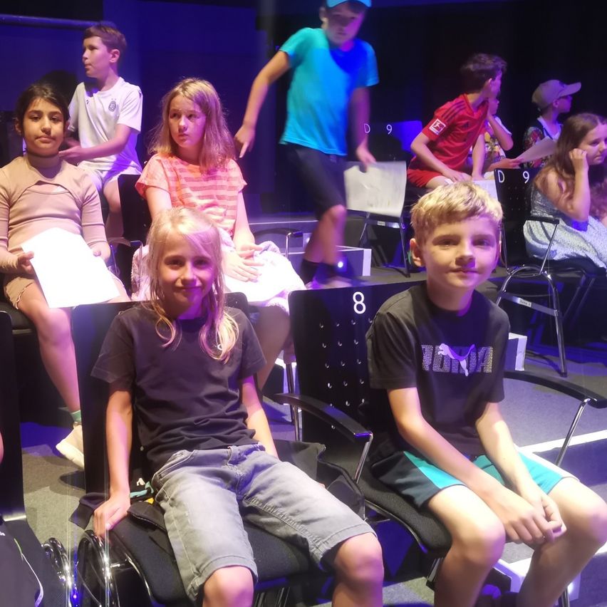 Kinder sitzen in Stühlen in einem Theater, ein Junge hält Papiere und ein anderer Junge lächelt. Auf der Bühne hinter ihnen sind blaue Lichter.