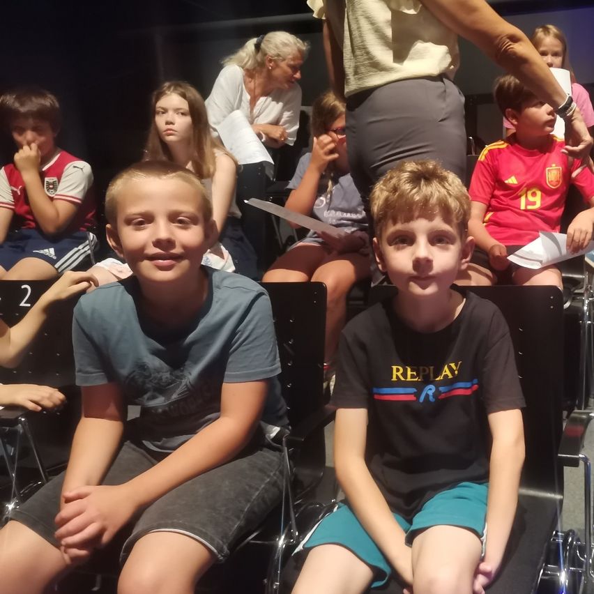 Mehrere Kinder sitzen in Reihen von Stühlen, einige in Sporttrikots. Zwei Jungen in der vorderen Reihe tragen blaue und schwarze T-Shirts. Dahinter hält eine Frau Papiere, und andere sitzen und beobachten.