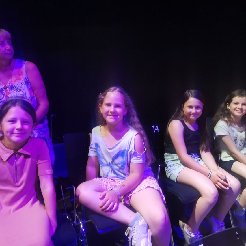 Vier Mädchen sitzen in Stühlen und lächeln in die Kamera. Eine trägt ein rosa Kleid, eine andere ein Schmetterlings-T-Shirt. Die Frau hinter ihnen schaut zu.