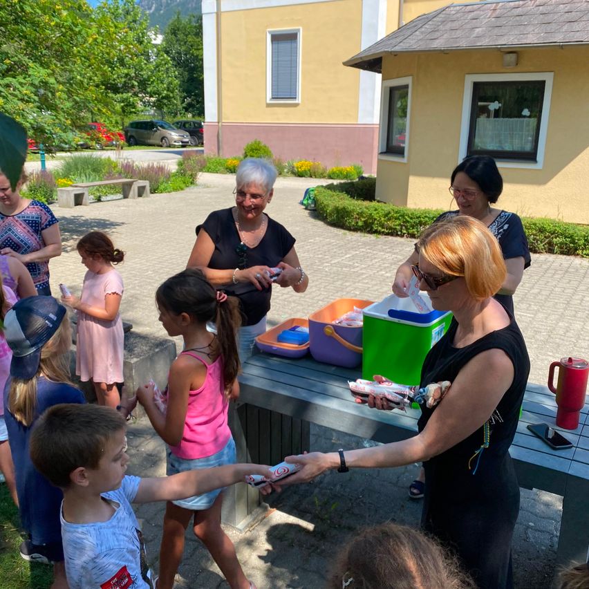 Erwachsene und Kinder versammeln sich um einen Tisch. Eine Frau überreicht einem Jungen ein Paket. Hinter ihr überreicht eine andere Frau einem Mädchen ein Paket. Der Ort ist im Freien, mit einem Gebäude im Hintergrund.