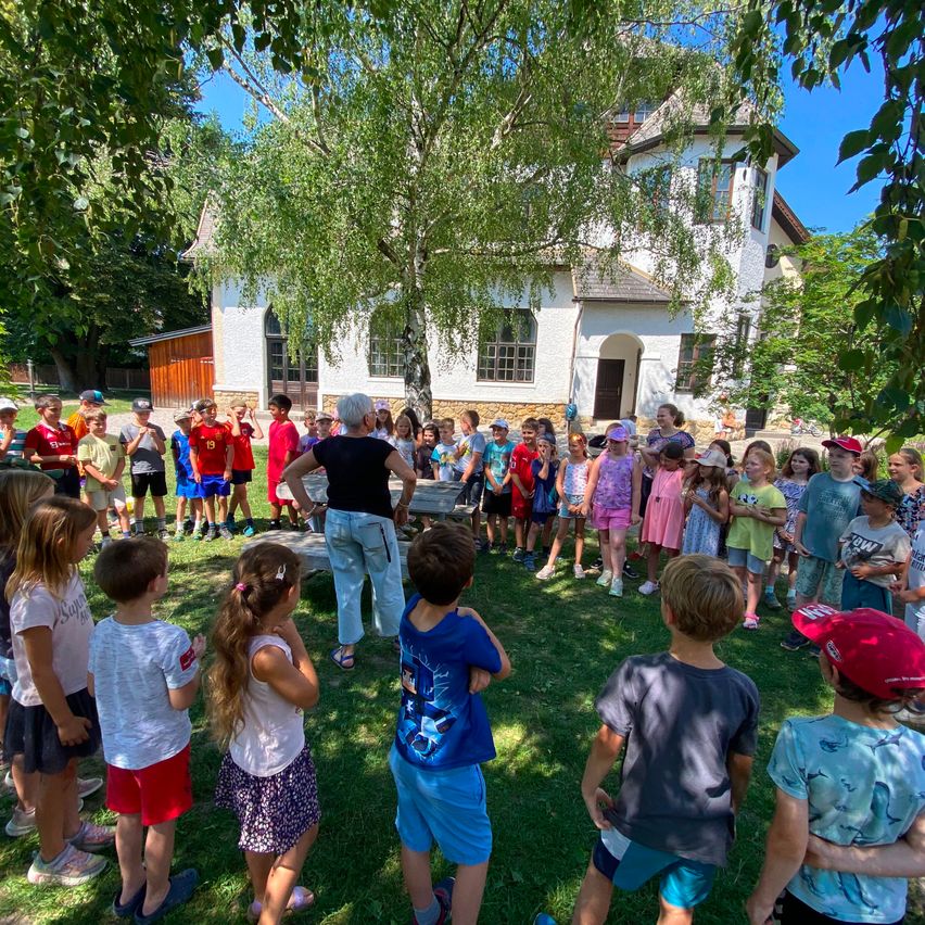 Eine Gruppe von Kindern und Erwachsenen versammelt sich vor einem weißen Haus, wobei eine Frau ein Gespräch unter einem Baum führt.