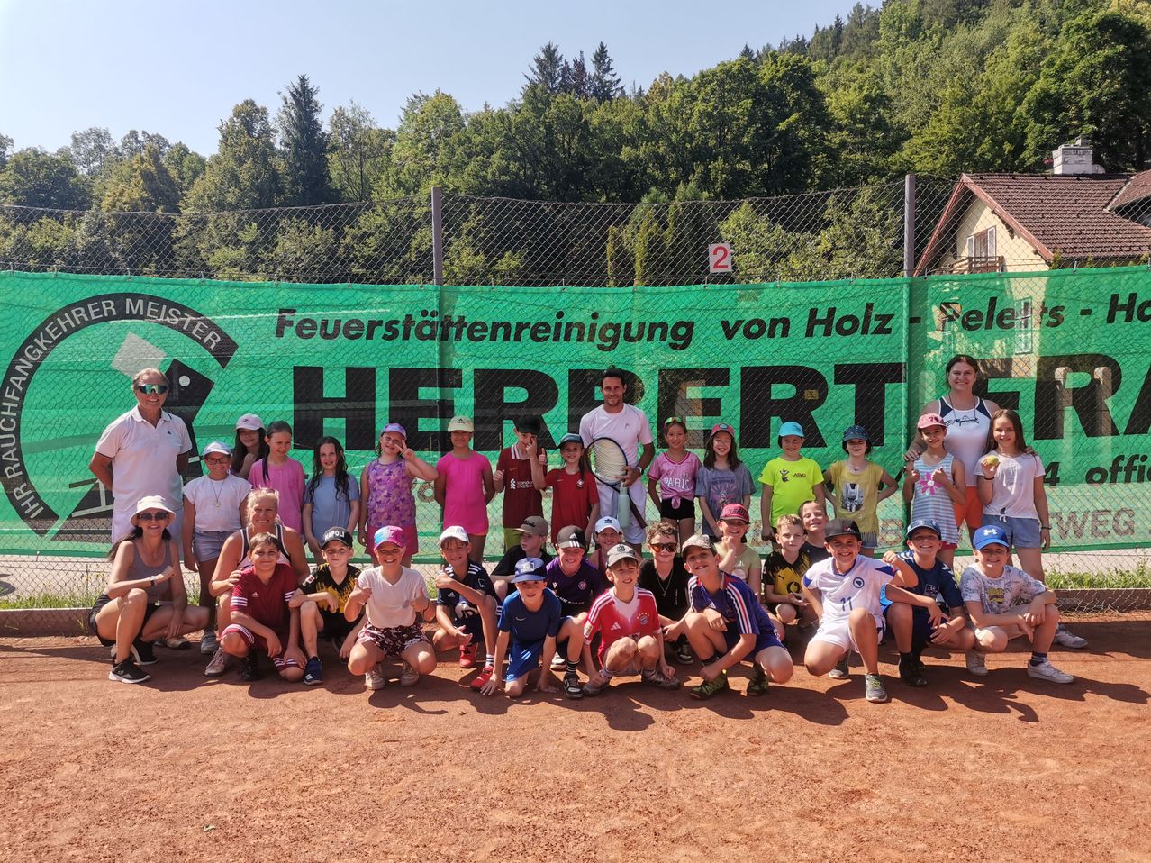 Eine Gruppe von Kindern und Erwachsenen posiert für ein Foto auf einem Tennisplatz. Im Hintergrund befindet sich ein grüner Banner mit Text und Logo. Hinter dem Zaun sind Bäume und ein Haus sichtbar.
