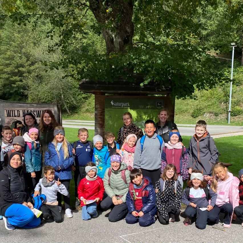 Eine Gruppe von Kindern und Erwachsenen posiert für ein Foto vor einem Schild in einem Park. Im Hintergrund sind Bäume und Grünflächen zu sehen.