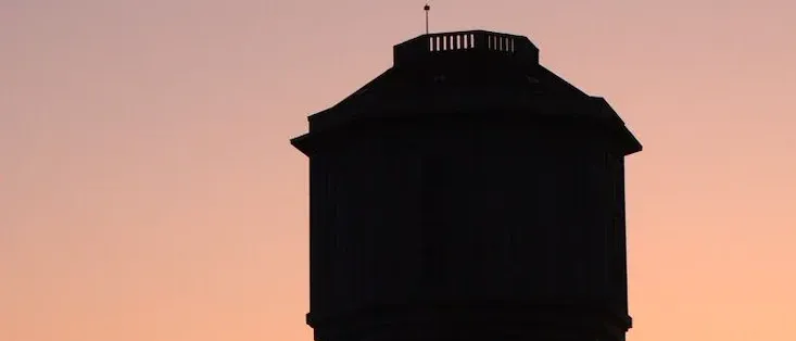 Die Silhouette eines Wasserturms steht vor einem Sonnenuntergangshimmel, mit Bäumen darunter sichtbar.