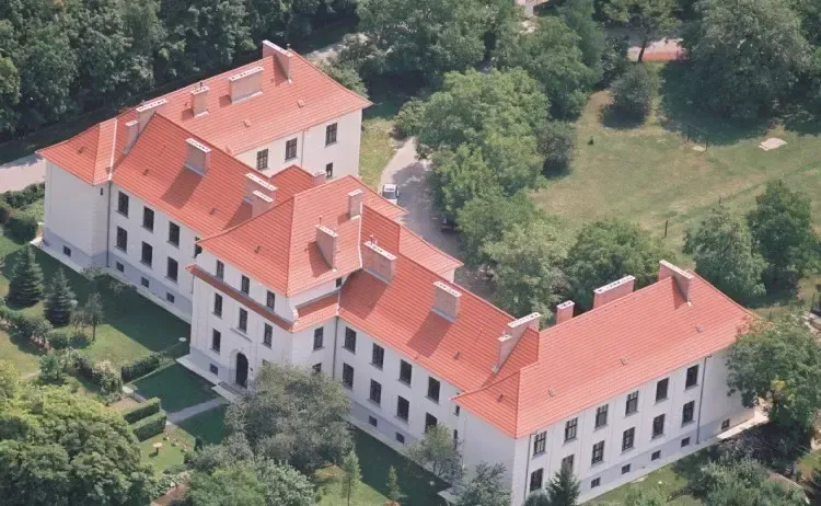 Luftaufnahme eines großen weißen Gebäudes mit roten Dächern und vielen Fenstern, umgeben von üppigem grünem Baumbestand.