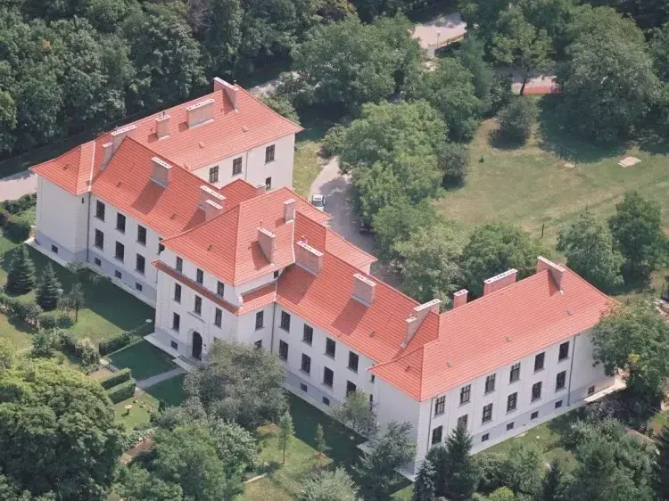 Luftaufnahme eines großen weißen Gebäudes mit roten Dächern und vielen Fenstern, umgeben von üppigem grünem Baumbestand.
