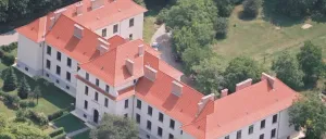 Luftaufnahme eines großen weißen Gebäudes mit roten Dächern und vielen Fenstern, umgeben von üppigem grünem Baumbestand.