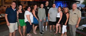 Eine Gruppe von Menschen steht nachts vor einer Bar und posiert für ein Foto. Die Bar hat Lichterketten und eine Bühne mit einer blauen Leinwand.