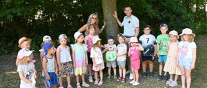 Eine Gruppe von Kindern und zwei Erwachsenen steht vor einem Baum in einem Wald. Die Erwachsenen lächeln, während die Kinder verschiedene Kleidungsstücke tragen und einen grünen Kreis halten.