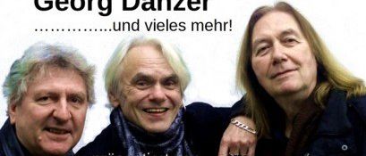 Drei Männer posieren für ein Foto vor weißem Hintergrund. Der Mann in der Mitte trägt einen Schal und lächelt. Der Mann auf der linken Seite hat graue Haare und trägt einen schwarzen Mantel. Der Mann auf der rechten Seite hat blonde Haare und trägt einen schwarzen Mantel. Der Text oben lautet Georg Danzer.