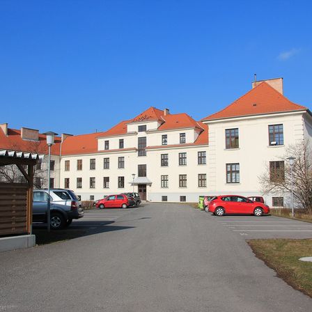 Ein großes weißes Gebäude mit roten Dächern und Fenstern befindet sich in einem Parkplatz, vor dem mehrere Autos geparkt sind.