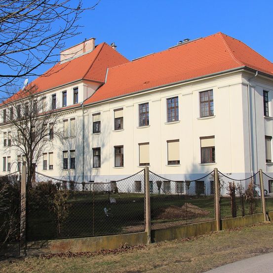 Ein großes weißes Gebäude mit rotem Dach und vielen Fenstern ist von einem Maschendrahtzaun umgeben. Der Zaun steht vor einem Grasbereich mit einigen Pflanzen.