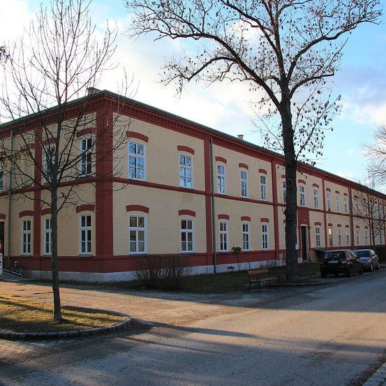 Ein Gebäude mit mehreren Fenstern und rotem Rand, mit zwei geparkten Autos davor. Bäume und eine Bank sind ebenfalls vorhanden.