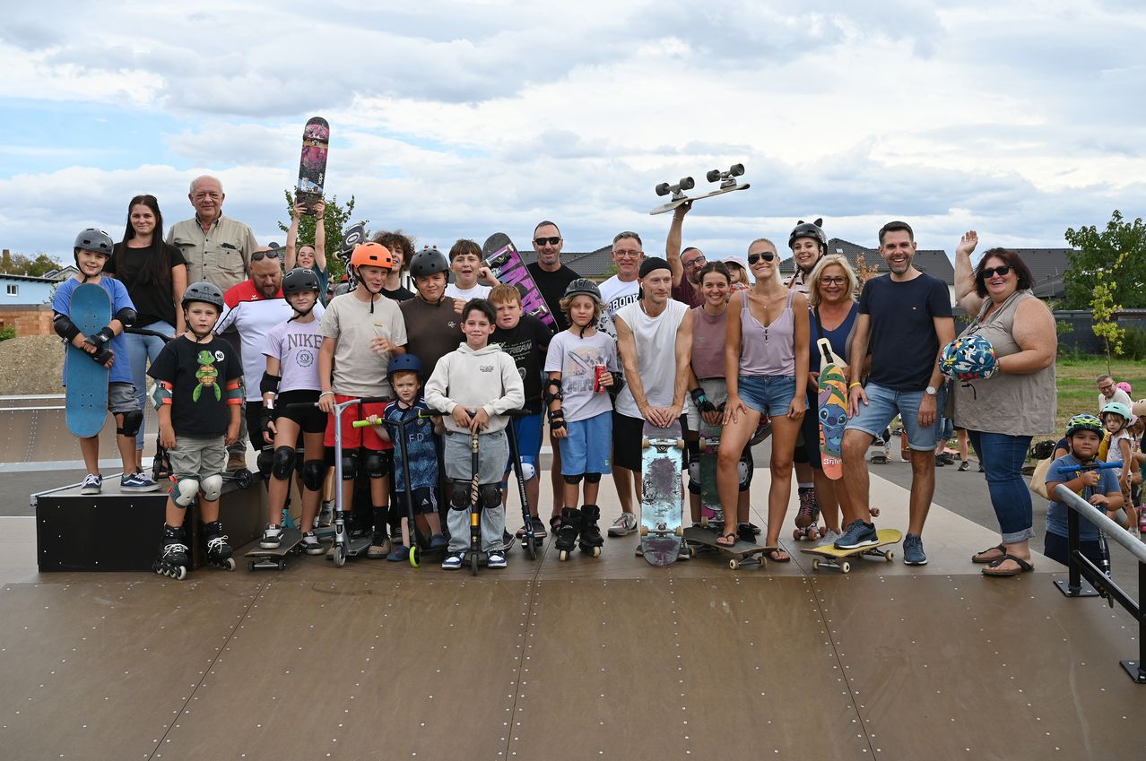 A group of people, including adults and children, are standing on a skate park with their skateboards and helmets. They are all smiling and posing for a photo.