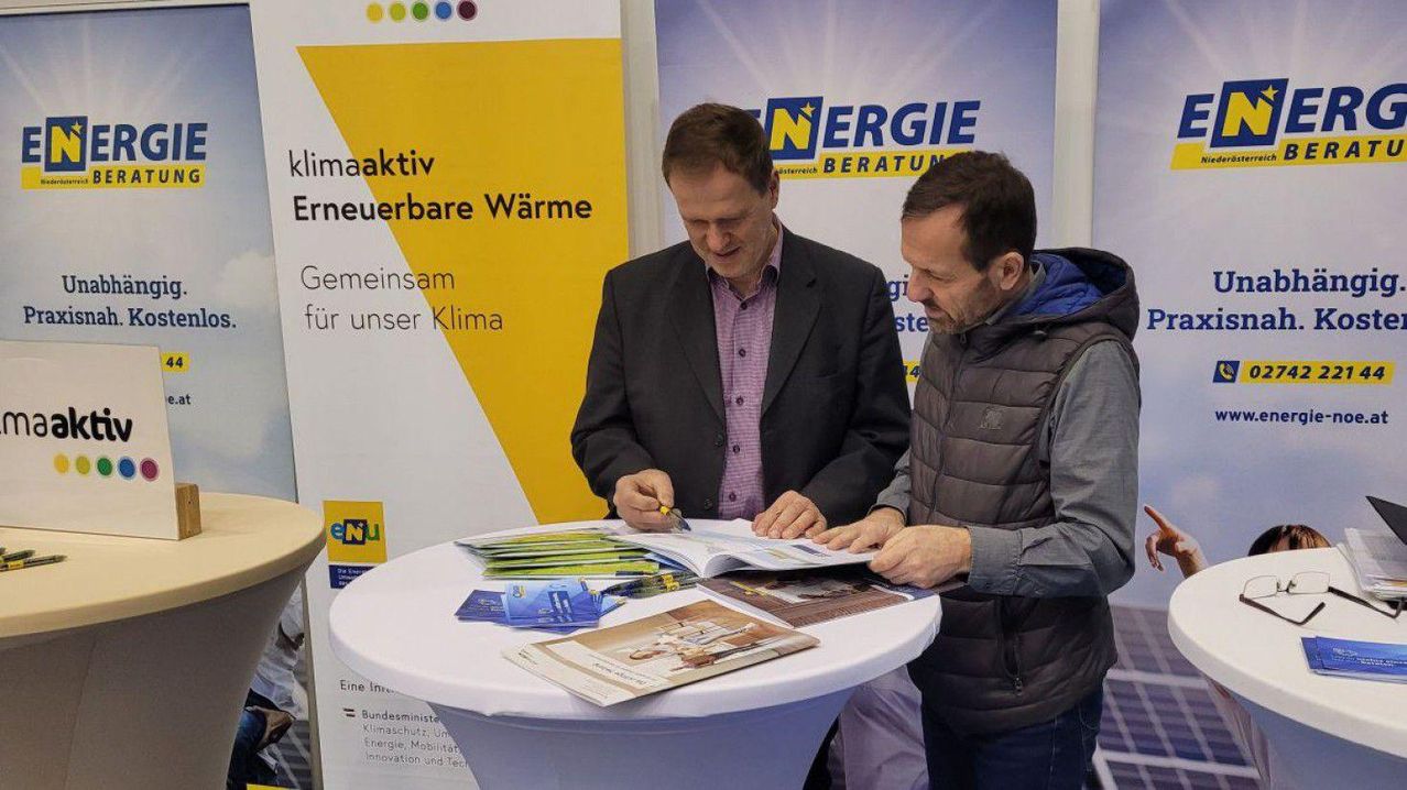 Two men at a booth, one signing a document, another reviewing pamphlets on a table. Behind them is a banner with 'ENERGIE' in blue and white.