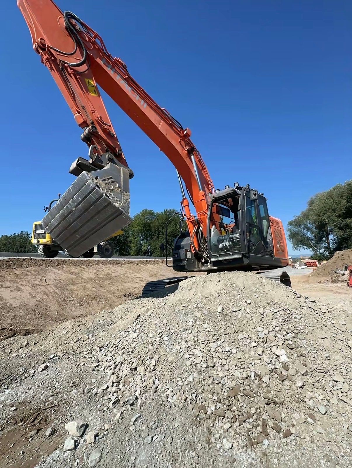 An orange excavator is scooping up a large pile of gravel on a sunny day at a construction site.