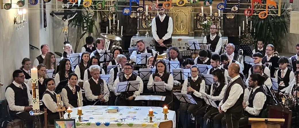 Ein Chor tritt in einer Kirche auf, vor einem Tisch mit weißer Tischdecke und Musiknoten. Der Chor ist von Musikinstrumenten umgeben und wird von Lampen beleuchtet.
