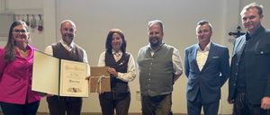 Fünf Personen stehen in einem Raum, lächeln und posieren für ein Foto. Eine Person hält ein Zertifikat. Sie tragen alle traditionelle Kleidung.