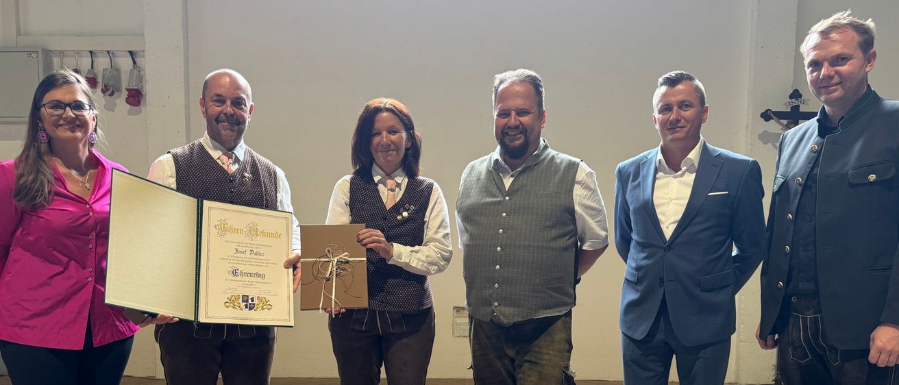 Fünf Personen stehen in einem Raum, lächeln und posieren für ein Foto. Eine Person hält ein Zertifikat. Sie tragen alle traditionelle Kleidung.