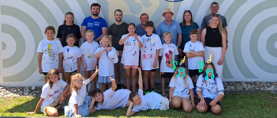Eine Gruppe von Menschen vor einer Wand mit einem Wappen, posiert für ein Foto. Einige stehen, andere knien und tragen verschiedene Shirts. Musiknoten wurden einigen Shirts hinzugefügt.