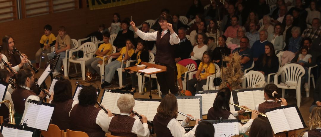 Ein großes Orchester spielt auf der Bühne, während ein Publikum zusieht. Die Musiker sitzen und spielen verschiedene Instrumente, während ein Dirigent vorne steht und die Aufführung leitet.