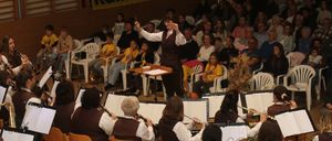 Ein großes Orchester spielt auf der Bühne, während ein Publikum zusieht. Die Musiker sitzen und spielen verschiedene Instrumente, während ein Dirigent vorne steht und die Aufführung leitet.