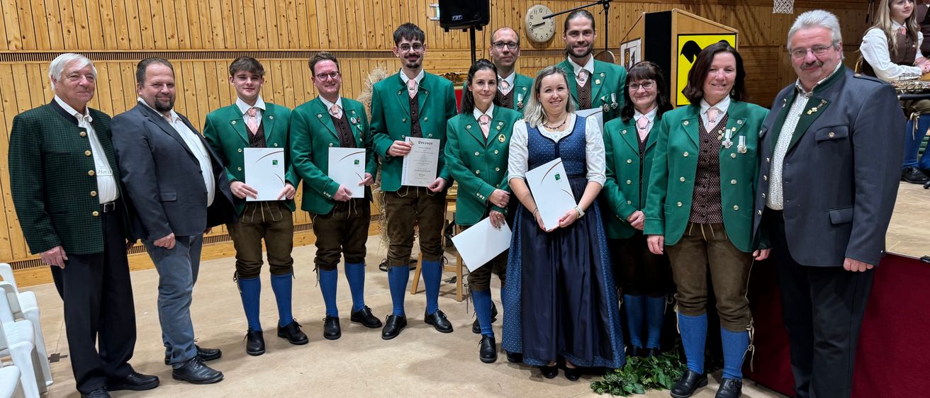 Eine Gruppe von Männern und Frauen in traditioneller Kleidung posiert für ein Foto und hält Urkunden in den Händen. Sie sind in einer Turnhalle mit Holzwänden und einem Basketballkorb.