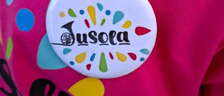 Ein rosafarbenes T-Shirt mit einem bunten Abzeichen, auf dem 'Anja' und 'Eusola' in der Mitte stehen, umgeben von verschiedenen Formen und Farben. Das Shirt hat zusätzlichen Text und Grafiken.