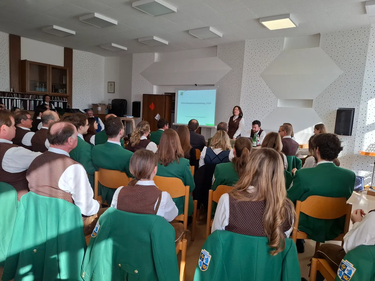 Eine Gruppe von Schülern in grünen Uniformen besucht eine Präsentation in einem Klassenzimmer mit einer Projektionsleinwand. Eine Frau steht vor dem Publikum, und einige Schüler sitzen mit Flaschen.