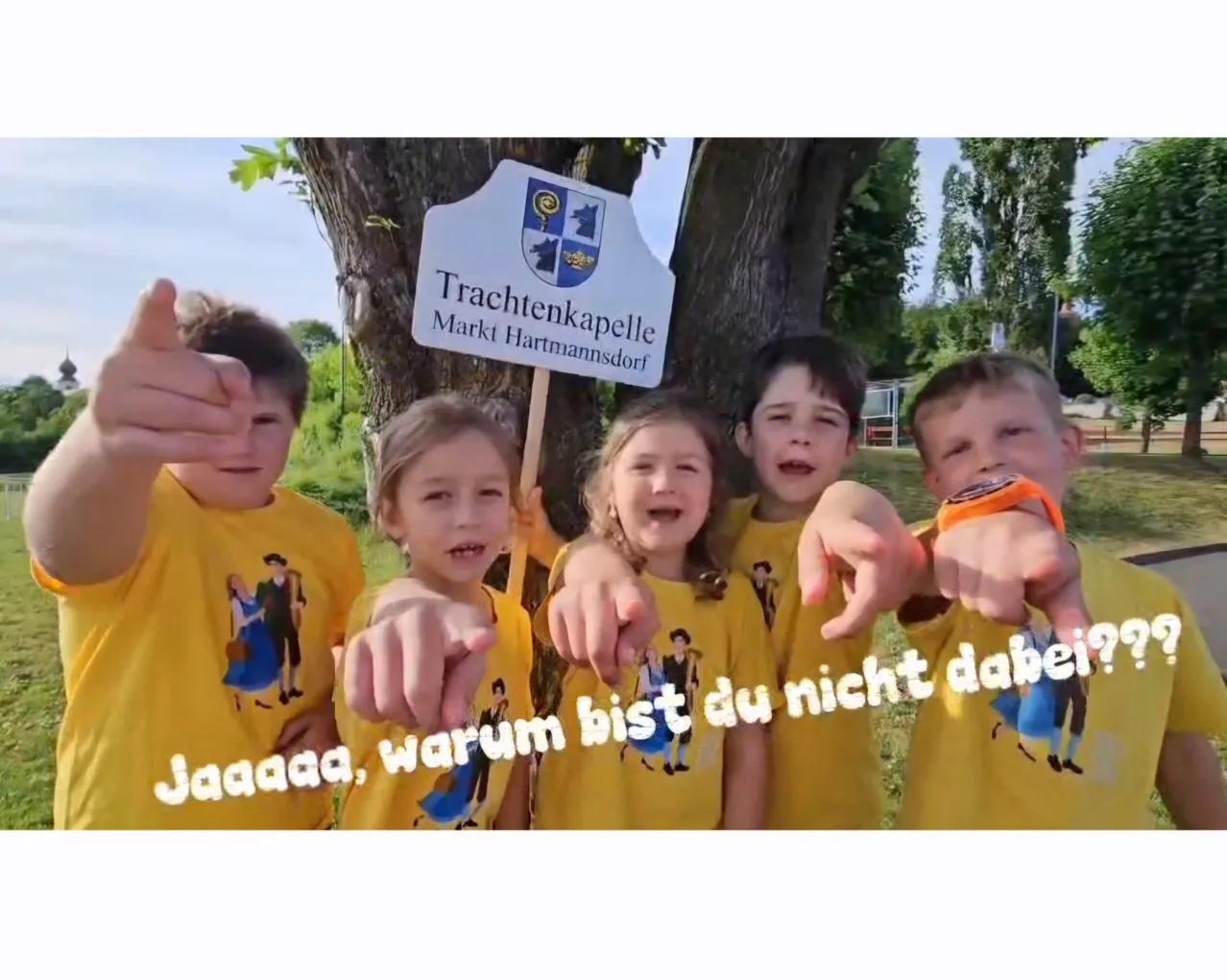 Eine Gruppe von Kindern in gelben T-Shirts posiert für ein Foto mit einem Schild, das 'Trachtenkappe' und 'Markt Hartmannsdorf' zeigt, in der Nähe eines Baumes.