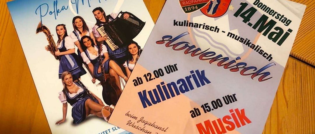 Zwei Poster sind auf einer Holzoberfläche zu sehen. Ein Poster zeigt eine Gruppe von Frauen, die Musikinstrumente spielen, mit dem Text 'Polka Madls'. Das andere Poster hat Text in Rot und Weiß, mit Veranstaltungsdetails wie 'Kulinarisch - Musikalisch', Datum '14. März' und Preisinformationen.