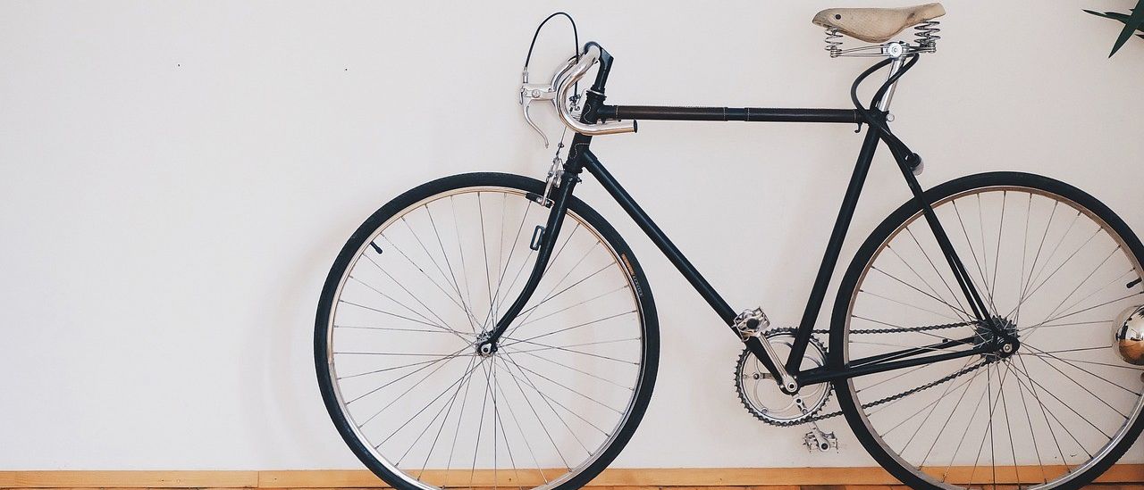 Ein schwarzes Fahrrad steht an einer weißen Wand gelehnt auf einem Holzboden. Das Fahrrad hat einen Holzsattel, eine Kette und Metalllenker.