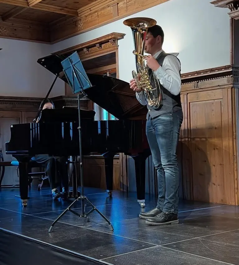 Ein Mann spielt ein Blasinstrument auf der Bühne, während ein anderer am Klavier sitzt. Der Raum ist mit Holzpaneelen geschmückt.